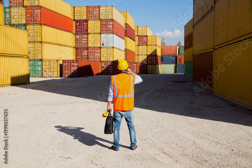 Construction worker in hard hat and safety vest operates radio at shipping container yard