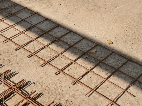  A rusty steel wire mesh grid lies on a sandy, light-colored concrete
surface at a construction site. A sharp diagonal shadow falls across the
industrial scene, creating a strong geometric contrast.