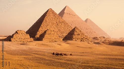 Group camels and their owners on desert plateau with Pyramids of Giza in soft morning light, Egypt. A group camels with their owners walks across sandy desert plateau near Pyramids of Giza at sunrise