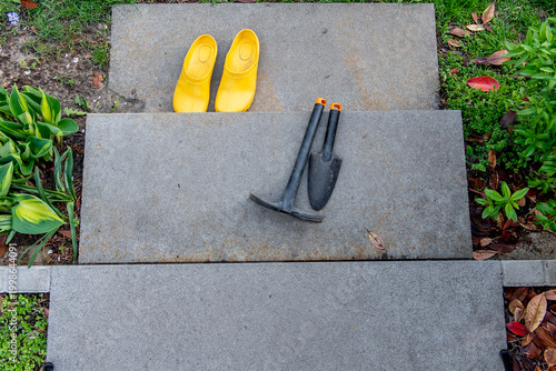 A pair of yellow garden clogs with a plastic trowel and a hoe on a concrete step in a garden.