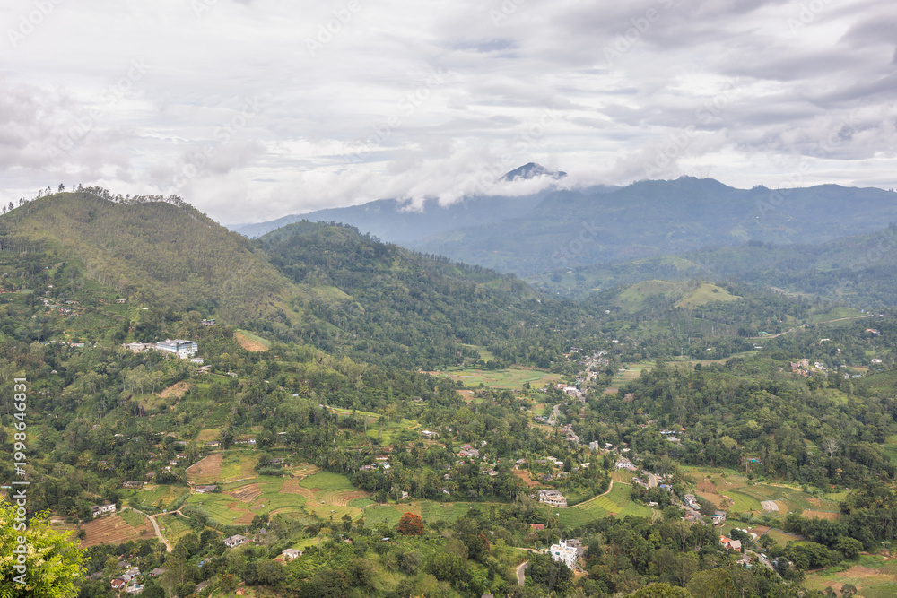Fototapeta premium Panoramic view of lush green mountain landscape with distant peaks shrouded in clouds under a dramatic sky. Mountain landscape near Ella town in Sri Lanka island in Asia.