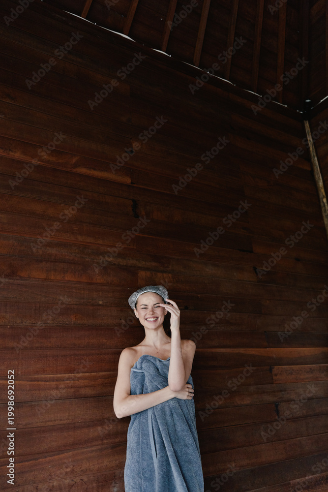 Fototapeta premium smiling woman wrapped in towel with hair wrap standing against wooden wall, relaxed and fresh indoors, wellness and spa concept