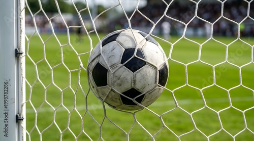Classic black and white soccer ball hitting the back of a white nylon goal net during a match, seen from behind the goal with a blurred stadium crowd and green grass pitch.