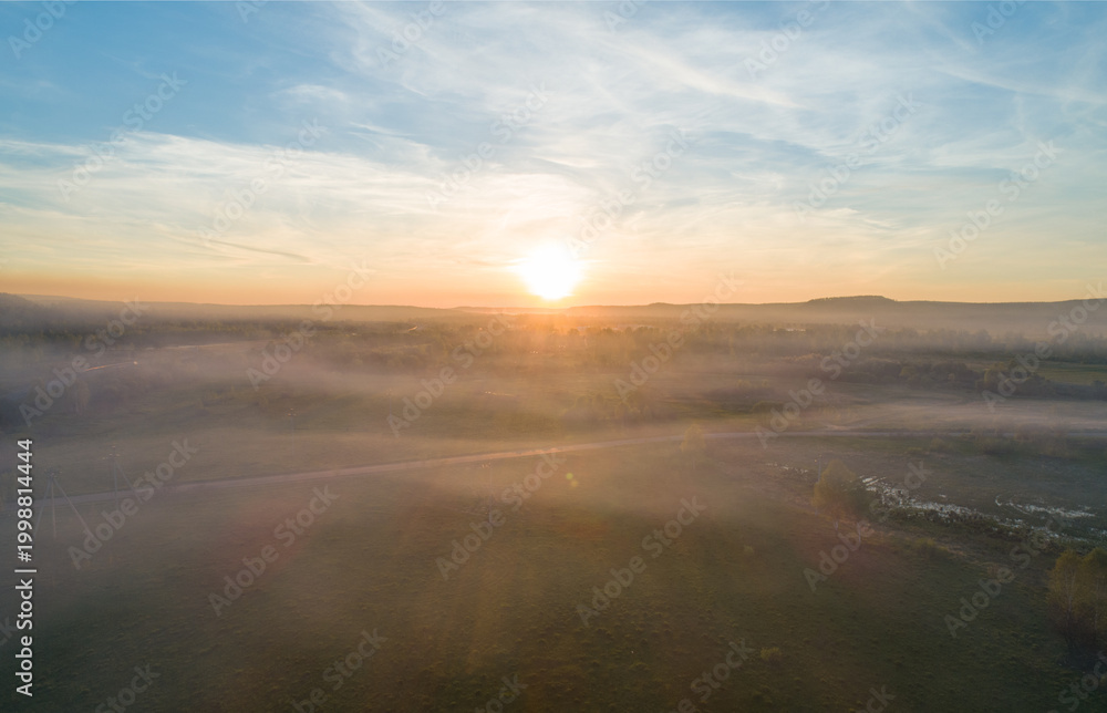 Fototapeta premium Aerial view captures bright sun cresting a misty valley landscape, casting warm light across green fields and distant mountains at dawn