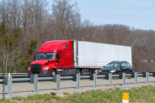 фотографія Bright red semi-truck with a white trailer drives down a highway alongside a black SUV, with a metal guardrail in the foreground and bare deciduous trees under a clear blue sky