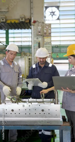 Asian factory team gathers near metal fixture, comparing tablet and laptop data while discussing inspection results, machine status, and production decisions on shop floor. Vertical