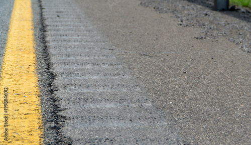 Photography Detailed close-up of a textured asphalt road surface with a bright yellow lane marking and a subtle rumble strip, illustrating infrastructure, wear, and travel