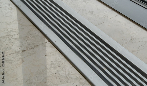 Billede på lærred Close-up of a textured anti-slip or drainage strip with parallel lines on a worn, cracked marble or concrete floor, creating a stark geometric pattern with contrasting shadows