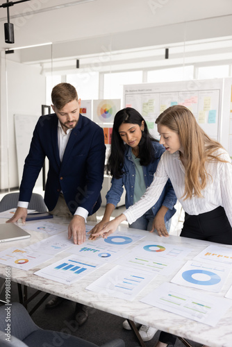 фотографія Three project managers engaged in teamwork in boardroom, reviewing business report, preparing for client presentation, discuss marketing strategy gather at desk with infographic data papers