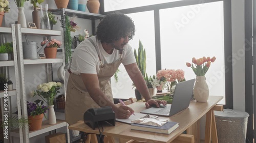 Man florist in apron leans over table typing on laptop and writes with pen in building; calm concentration.