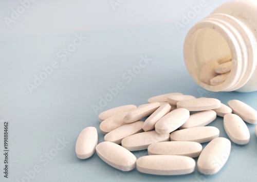 White oval pills are spilling from an open plastic medicine bottle onto a light blue surface