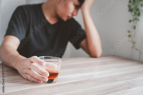 Foto Depressed asian man sitting at a wooden table holding a glass of whiskey, feeling stressed, and struggling with alcohol addiction