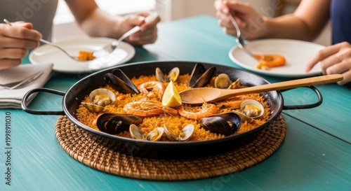 Paella seafood dish presentation in large pan on table with people eating in background.