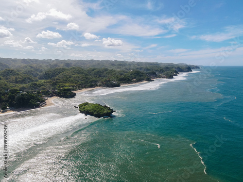Aerial view of Baron Beach with turquoise ocean waves crashing against a rocky outcrop and lush green hills under a bright sky Tanjungsari, Special Region of Yogyakarta, Indonesia.