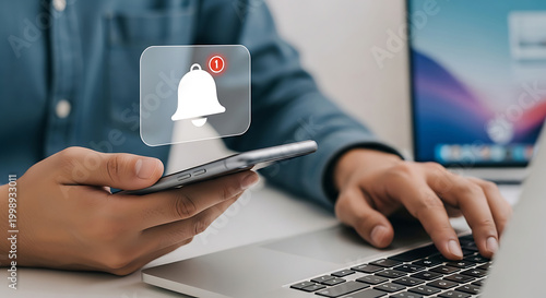 Person receiving notification alert on smartphone while working on laptop, digital technology concept