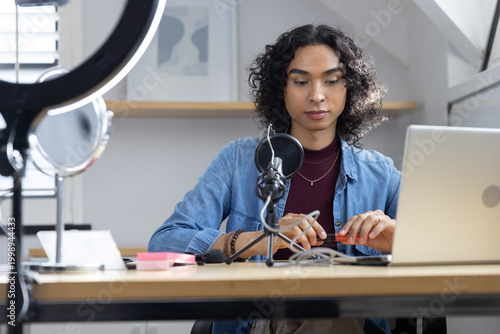 Non-binary adult adjusting microphone and handling small orange object at studio desk with laptop