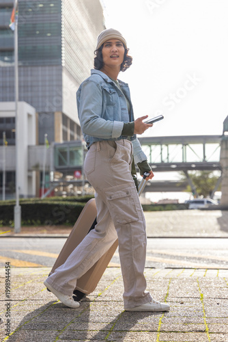 Woman in denim jacket walking across urban plaza pulling rolling suitcase and holding phone