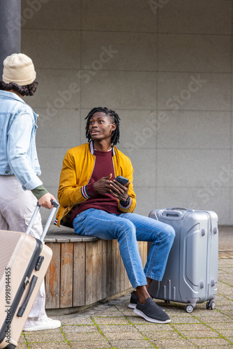 Travel companions sitting on bench wearing yellow jacket, holding phone, moving suitcase at station