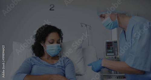 Listening patient in blue top mask on hospital bed, nurse in scrubs gesturing by IV pump