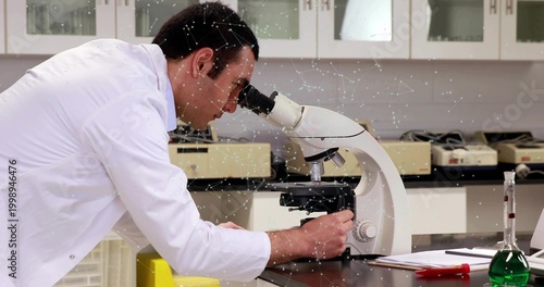 Leaning scientist in white lab coat peering into microscope at research lab, green flask nearby