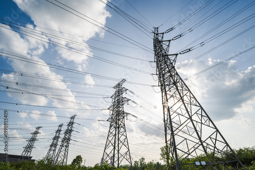 Fotografía This image shows multiple steel lattice high voltage transmission pylons strung with power lines, set against a bright cloudy daytime sky with green vegetation at the base