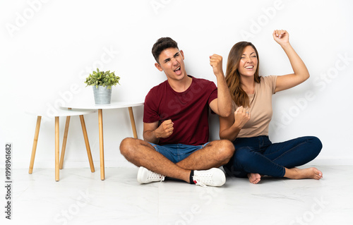 Fotografie Young couple sitting on the floor isolated on white background celebrating a vic