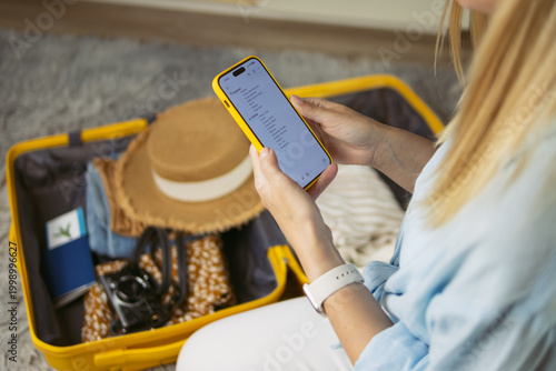 фотографія Woman checking travel checklist on smartphone, preparing suitcase for summer vac