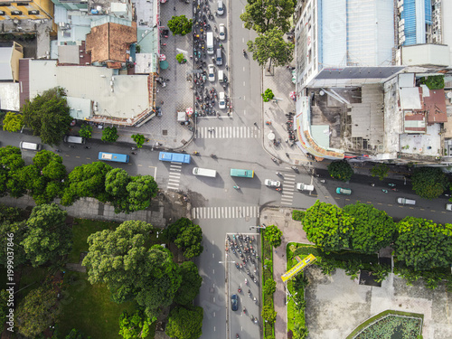 Aerial view of a busy urban intersection with heavy motorbike traffic, colorful buses, and lush green trees lining the streets Ho Chi Minh City, Ho Chi Minh, Vietnam.