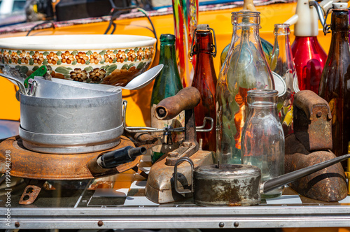 Assorted vintage antiques and flea market finds including rusted flat irons, various glass bottles, an aluminum pot with a spoon, and a floral ceramic bowl displayed in a sunny outdoor setting.