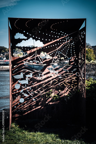 Description (191 characters):
Rusty Corten steel outdoor art sculpture with laser-cut coastal scene of boats, birds and waves, standing beside a river estuary under a blue sky.