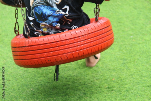 A creative upcycled tire swing painted bright orange on green artificial turf at a playground. Concept of eco-friendly recreation, sustainable DIY play equipment, and outdoor childhood fun.