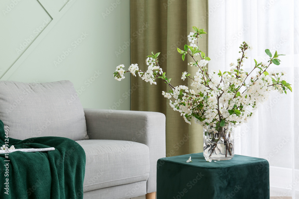 Fototapeta premium Interior of light living room with blossoming branches in vase, sofa and pouf