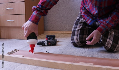 A man in a plaid suit sits on the floor in a bedroom and applies glue from a tube to the inside of a wooden bed's side plank, DIY home furniture repair