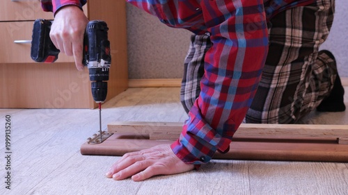 A worker in a plaid shirt operates a screwdriver on a disassembled wooden bed on a light floor in a home room, fixing fasteners on the side board of a light wood bed
