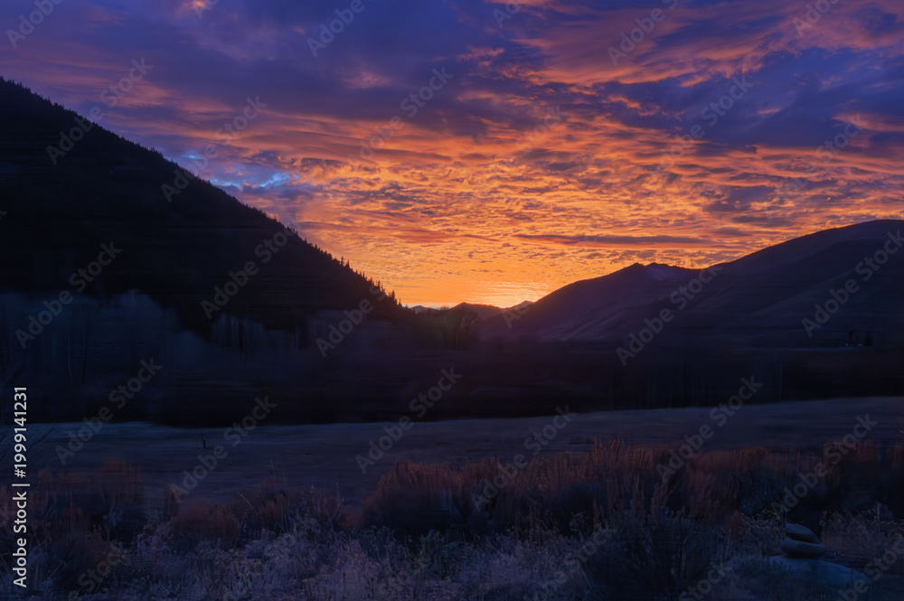 Obraz premium Majestic mountains frame a fiery sunset sky. Valley floor glows with soft, silvery frost. Deep purples blend with orange hues in clouds. Silent, frost-kissed grasses dot the foreground