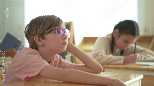 Schoolboy looking bored and distracted in classroom while classmates study, representing lack of focus and attention