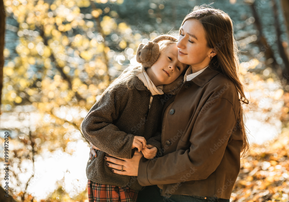 Fototapeta premium Heartwarming moment: loving mother hugging her little daughter wearing earmuffs in beautiful autumn forest.