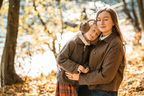 Heartwarming moment: loving mother hugging her little daughter wearing earmuffs in beautiful autumn forest.