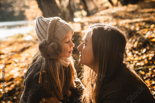 Happy mother and little daughter smiling at each other in autumn forest with golden leaves background.