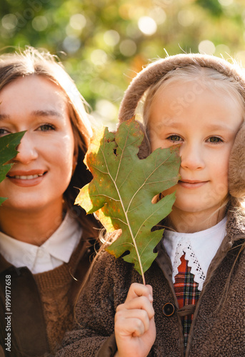 Creative autumn portrait: mother and daughter looking at camera with golden oak leaves covering half of their faces.