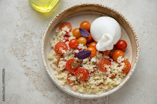 Bowl with couscous, cherry tomatoes and burrata cheese on a light-beige stone background, horizontal shot, elevated view