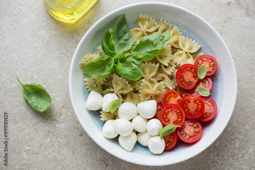 Plate of caprese pasta salad with mini mozzarella, cherry tomatoes and farfalle, horizontal shot on a beige stone background, high angle view