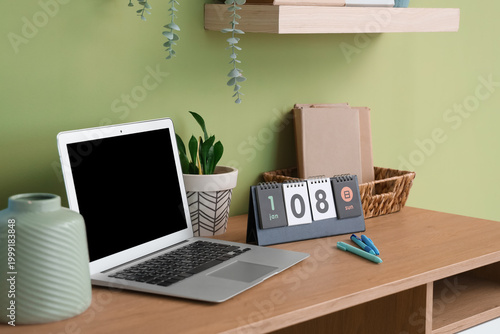 Modern laptop, calendar, books and pens on wooden table near color wall