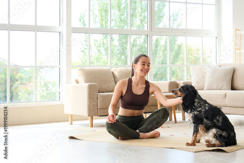 Sporty young woman with cute dog sitting on yoga mat at home
