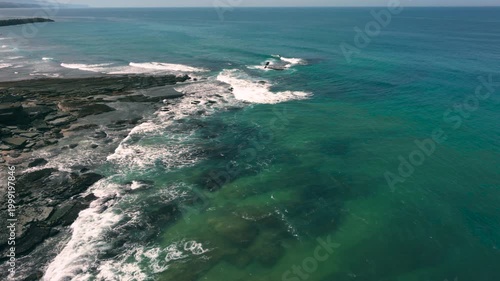Aerial shot of the ocean with waves doing swirls and patters, rocks, sand and beach