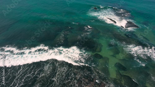 Aerial shot of the ocean with waves doing swirls and patters, rocks, sand and beach