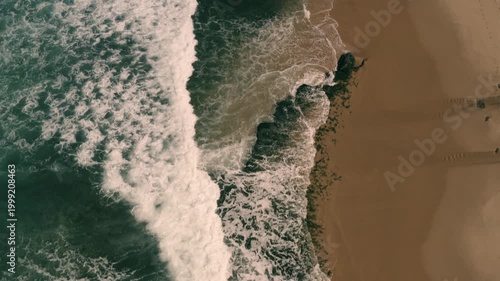 Aerial shot of the ocean with waves doing swirls and patters, rocks, sand and beach