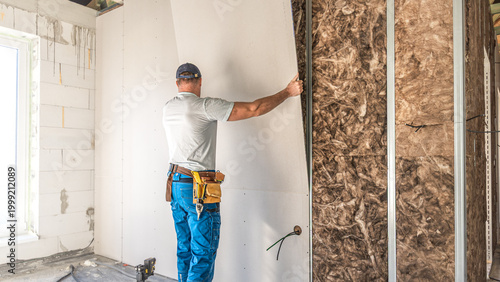 Making a plasterboard partition. A worker is laying drywall board on a metal support structure of partition.