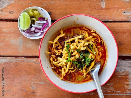 Traditional Northern Thai curry noodle soup (Khao Soy) with tender beef, topped with crispy noodles. Served with lime, red onions, and pickled cabbage on a wooden table.