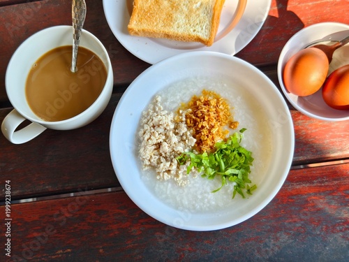 Savory homemade Asian breakfast. Rice congee with minced pork, ginger, and fried shallots. Served with hot milk coffee, toast, and two hard-boiled eggs on a wooden table with morning light.